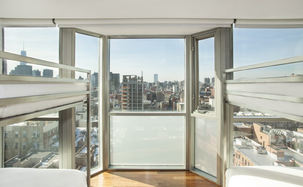 A room with a city view, featuring a bed and chair in front of large windows.